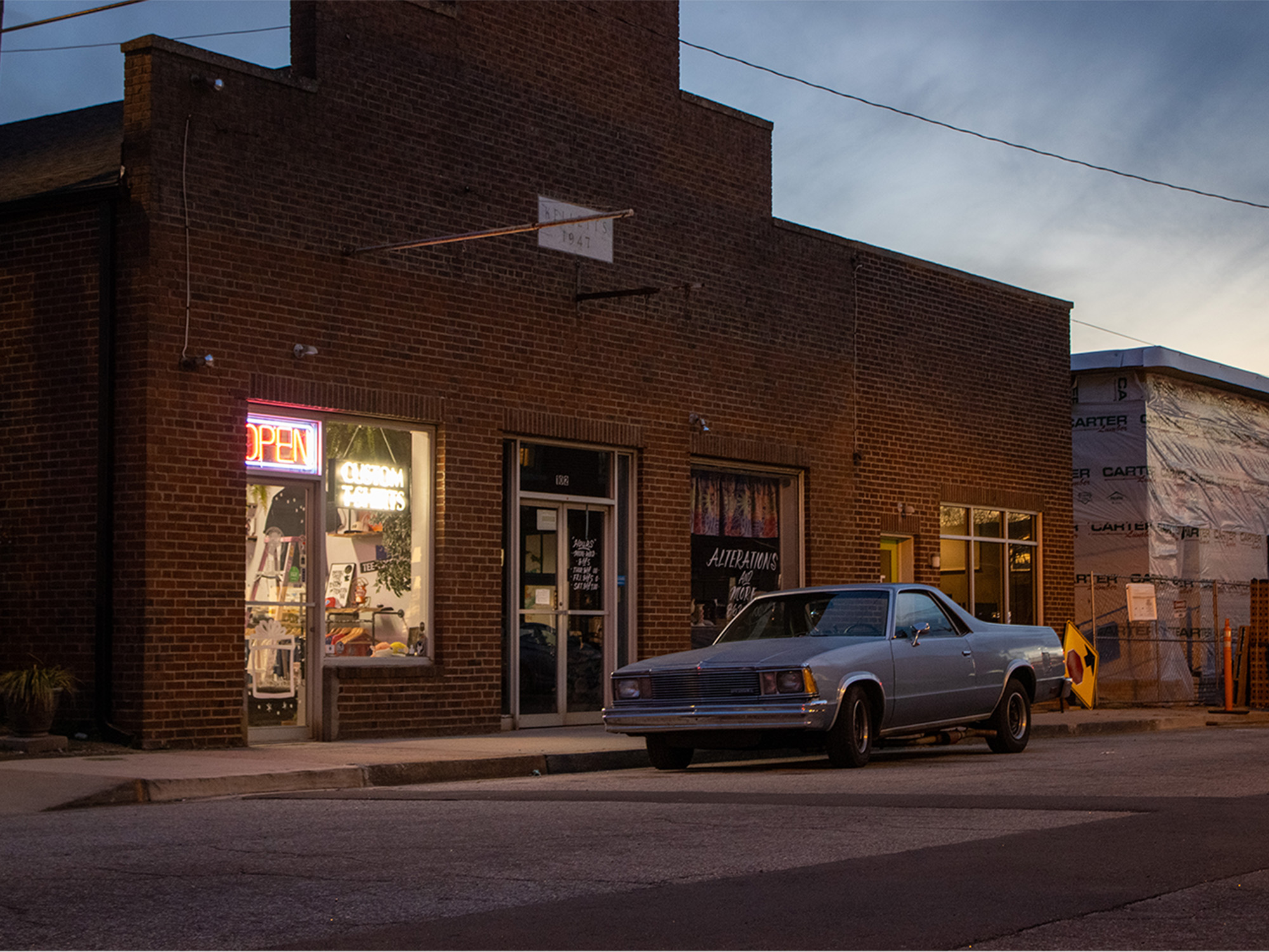 el camino parked on street at sunset in front of brick building with neon open sign in window